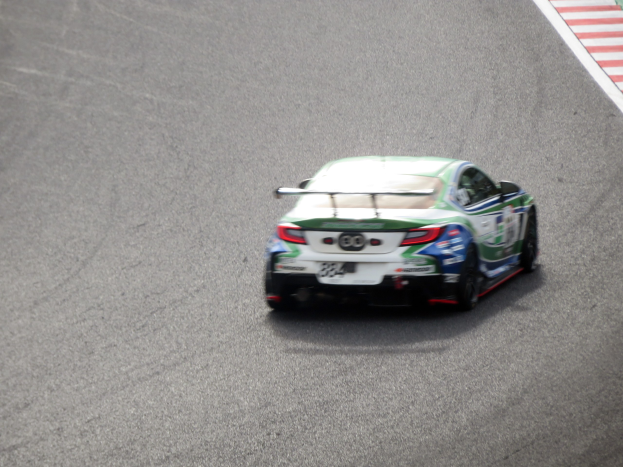 BMW M4 GT3 Rennauto auf einer Rennstrecke mit einem Rad im Vordergrund und einer textbehafteten Wand auf der rechten Seite.