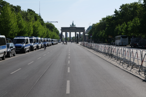 Eine Reihe von Polizeiwagen, die auf der Seite einer Straße vor dem Brandenburger Tor in Berlin, Deutschland, geparkt sind, mit Menschen, die Fahrräder fahren und auf der Straße stehen, Barrieren, Bäumen und einem Bogen mit Statuen im Hintergrund.
