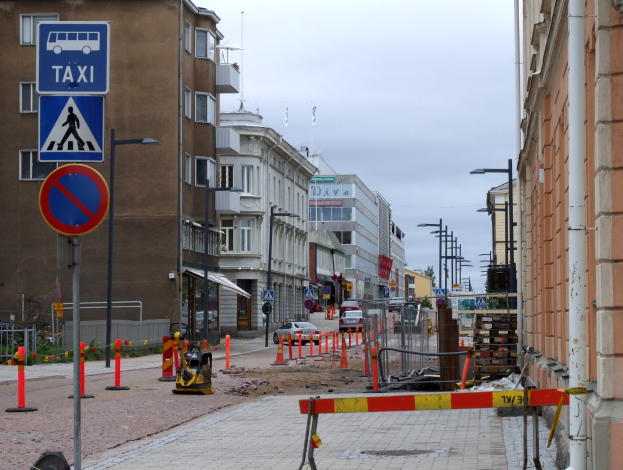 Eine Stadtstraße mit Gebäuden, Straßenschildern, Straßenlaternen, Verkehrszeichen, Verkehrskegeln, Kraftfahrzeugen, Absperrpollern, Bäumen und einem Himmel mit Wolken im Hintergrund.