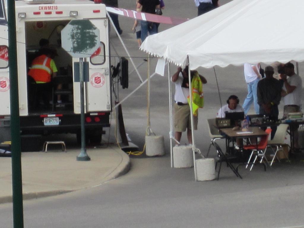 Ein Krankenwagen steht links auf der Straße, während rechts ein Zelt viele Menschen schützt, mit einem Tisch und einem Stuhl in der Nähe.