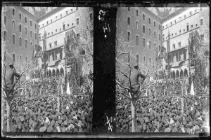 Ein Schwarz-Weiß-Foto einer großen Menge, die vor einem mehrstöckigen Gebäude mit Fenstern, Bäumen und Fahnen im Hintergrund protestiert, einige halten Schilder und Banner.