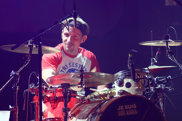 Ein Mann spielt Schlagzeug auf der Bühne bei einem Konzert, trägt ein rotes und weißes T-Shirt und lächelt, mit mehreren Mikrofonen vor ihm.