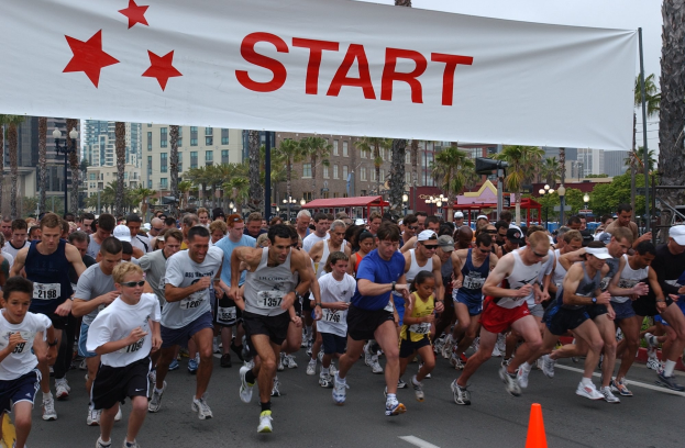 Gruppe von Läufern bei einem Marathon, die an einer Verkehrskegel vorbei laufen, mit einem Banner und Gebäuden im Hintergrund bei klarem blauem Himmel.