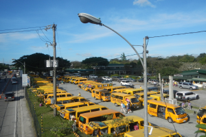 Eine große Anzahl gelber Schulbusse, die an einer Straße geparkt sind, mit Fußgängern auf dem Gehweg, Strommasten, Bäumen, Gebäuden und einem bewölkten Himmel im Hintergrund.