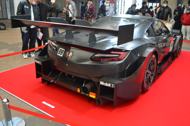 Ein roter und weißer Honda NSX GT3 Rennauto auf der Tokyo Motor Show, umgeben von Menschen in verschiedenen Kleidern und einigen mit Masken, mit einem roten Teppich daneben und Schildern und Lichtern im Hintergrund.