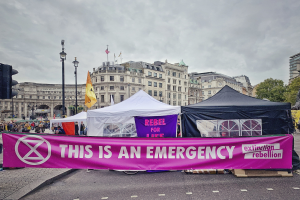 Gruppe von Menschen mit einer "Das ist ein Notfall"-Plakette in Trafalgar Square, London, mit Zelten, Fahnen und Gebäuden im Hintergrund.
