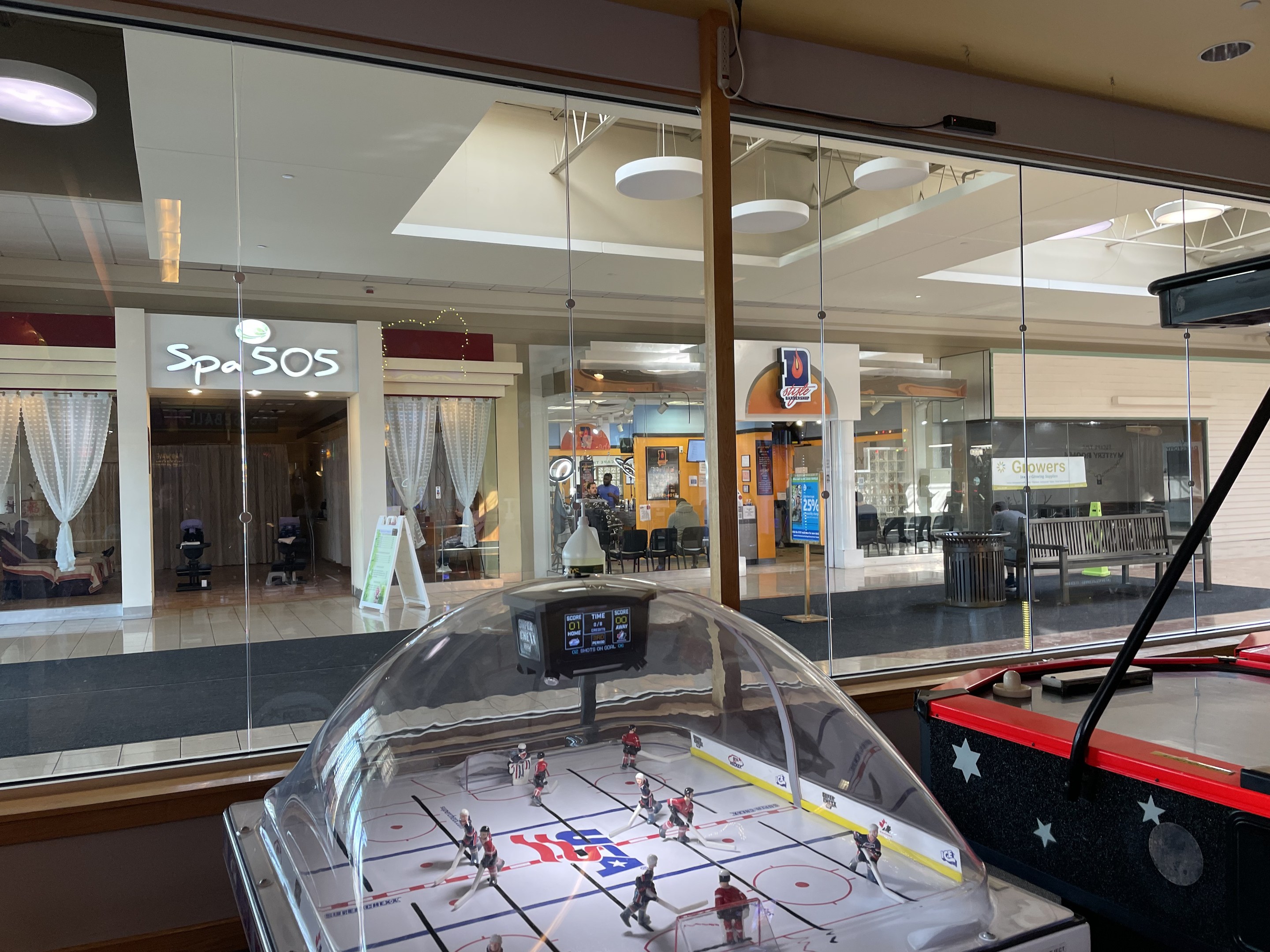 Eislaufbahn in einem Einkaufszentrum, umgeben von Glaswänden, mit einem Air-Hockey-Tisch auf der rechten Seite und verschiedenen Gegenständen, die durch das Glas sichtbar sind.
