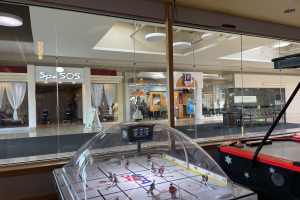 Eislaufbahn in einem Einkaufszentrum, umgeben von Glaswänden, mit einem Air-Hockey-Tisch auf der rechten Seite und verschiedenen Gegenständen, die durch das Glas sichtbar sind.