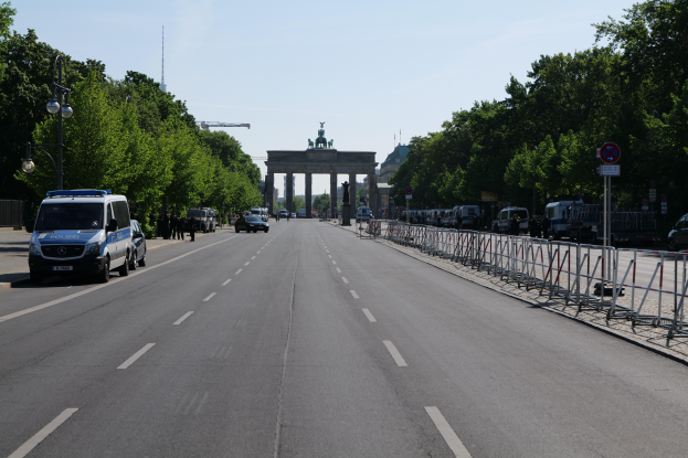 Ein Polizeiwagen steht auf der Seite einer vielbefahrenen Straße vor dem Brandenburg Tor in Berlin, Deutschland, mit Barrieren, Schildern, Bäumen, Laternenpfählen und einem bewölkten Himmel.
