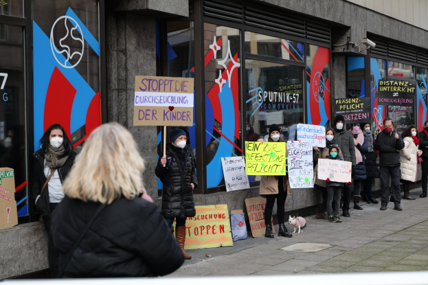 Maskierte Menschen mit Schildern und Plakaten protestieren vor einem Glasfrontgebäude, mit Kameras und einem Hund anwesend, wahrscheinlich gegen ein deutsches Schulmaskenverbot.