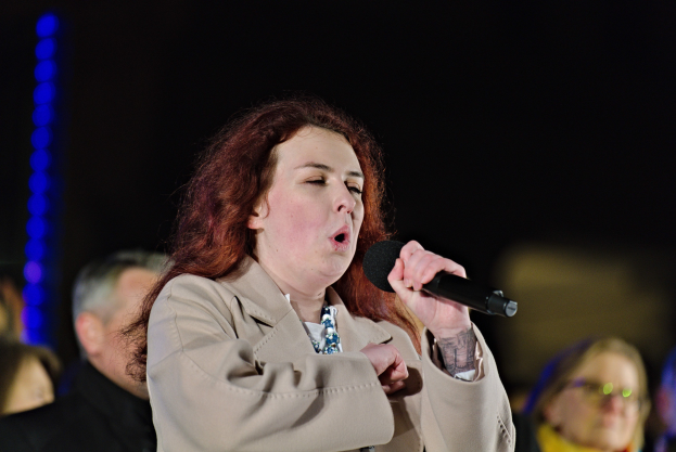 Eine Frau singt in ein Mikrofon auf der Bühne mit zwei Personen dahinter, unter Bühnenbeleuchtung.