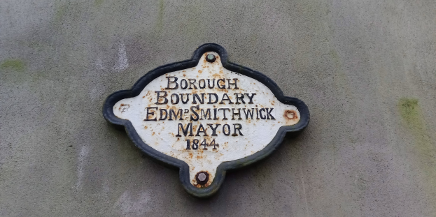 Metallplatte an einer Gebäudeseite, die die Stadtteilgrenze markiert, mit "Stadtteilgrenze Edm Smithwick Bürgermeister" in fetten weißen Buchstaben auf einem schwarzen Hintergrund.