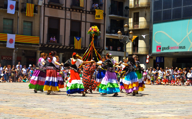 Eine Gruppe von Frauen in bunten Kleidern, die auf der Straße tanzen, umgeben von einer Menge Menschen, mit Gebäuden, Gelöndern, Balkonen, Flaggen, einem Lichtmast und einer Tafel mit einer Aufschrift im Hintergrund.