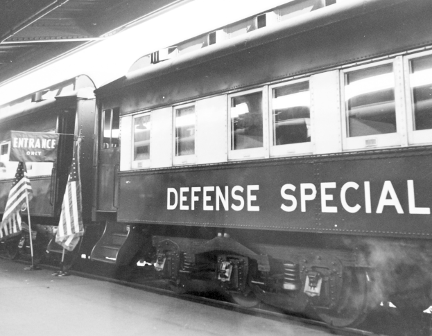 Schwarz-weißes Foto eines Zuges mit der Aufschrift "Defense Special" auf den Eisenbahnschienen mit zwei Fahnen links und einer Decke oben.