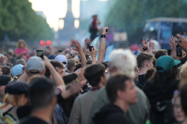 Eine Menschenmenge auf einem Musikfestival mit erhobenen Händen, einige tragen Käppchen und halten Mobiltelefone, mit unscharfen Bäumen, Gebäuden, Lichtern und einem Fahrzeug im Hintergrund.
