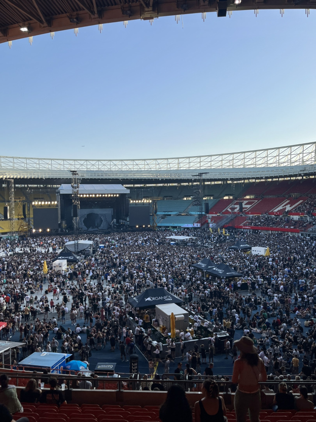 Große Menschenmenge auf Tribünen und vor der Bühne in einem Stadion mit Zelten, Geländern und Scheinwerfern unter einem Himmel, was auf ein Konzert in der O2 Arena in London hindeutet.