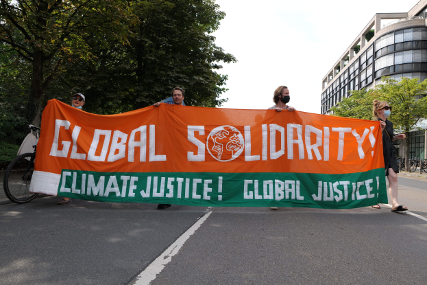 Menschen marschieren auf einer baumbestandenen Straße mit einem Banner "Globale Solidarität: Klimagerechtigkeit" und parkenden Fahrrädern sowie Gebäuden im Hintergrund.