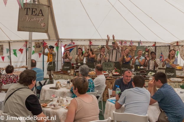 Eine Gruppe von Menschen sitzt an Tischen in einem Zelt, umgeben von Fahnen und einer Tafel mit Text, mit Tassen, Untertassen, Tellern, Gläsern und Flaschen auf den Tischen und anderen im Hintergrund stehend und klappend während eines Teeempfangs.