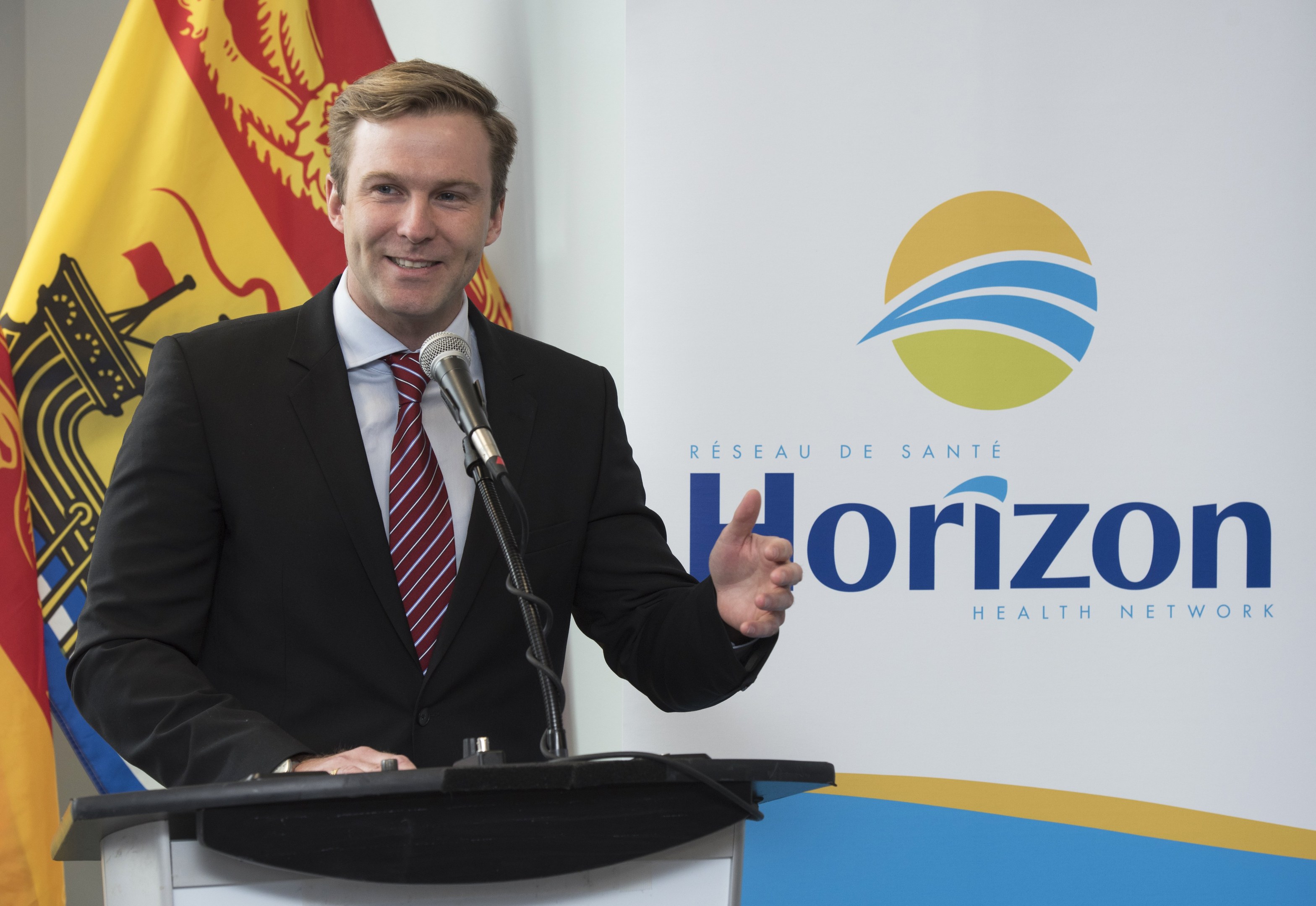 Ein Mann in einem schwarzen Blazer, weißem Hemd und roter Krawatte steht an einem Rednerpult mit einem Mikrofon und einem Banner im Hintergrund und lächelt.
