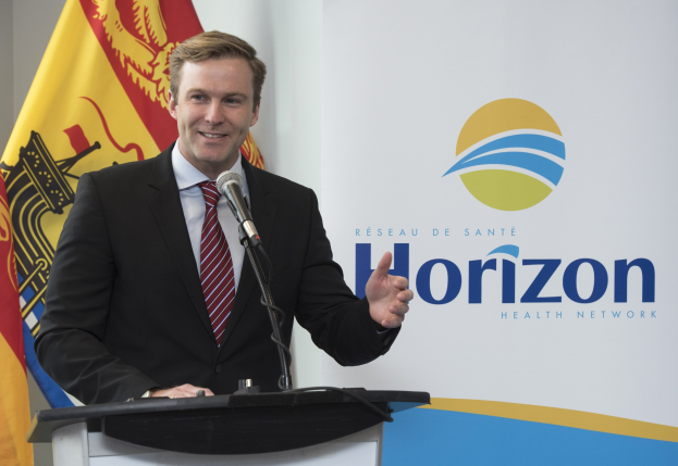 Ein Mann in einem schwarzen Blazer, weißem Hemd und roter Krawatte steht an einem Rednerpult mit einem Mikrofon und einem Banner im Hintergrund und lächelt.