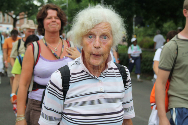 Eine öltere Frau mit öffentlicher Miene geht die Straße entlang, umgeben von Menschen, einige tragen Taschen, mit Bäumen, Gebäuden und einem klaren Himmel im Hintergrund.