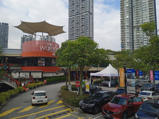 Eine belebte Stadtstraße mit parkenden Autos, hohen Gebäuden, Bäumen und Passanten, mit dem 'Waterfront Shopping Centre' auf der linken Seite und einer bewölkten Kulisse.