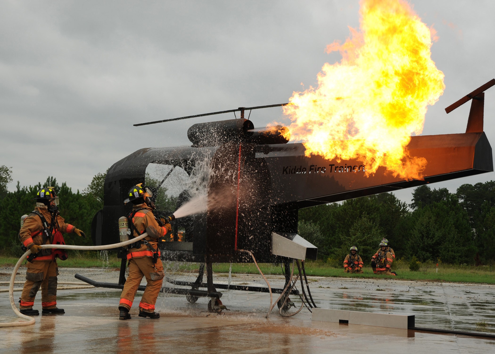Feuerwehrleute in Helmen spritzen Wasser auf einen brennenden Hubschrauber, während Bäume und ein blauer Himmel im Hintergrund zu sehen sind.