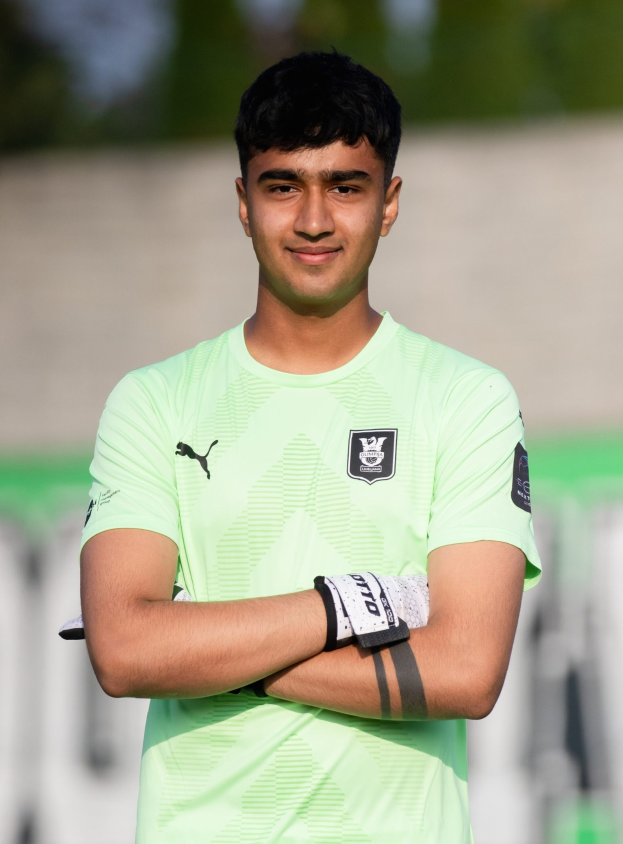Ein junger Mann in einem grünen T-Shirt steht auf einem Fußballfeld und hat die Arme verschränkt, er lächelt, wahrscheinlich ein Torwart, mit einem unscharfen Hintergrund.