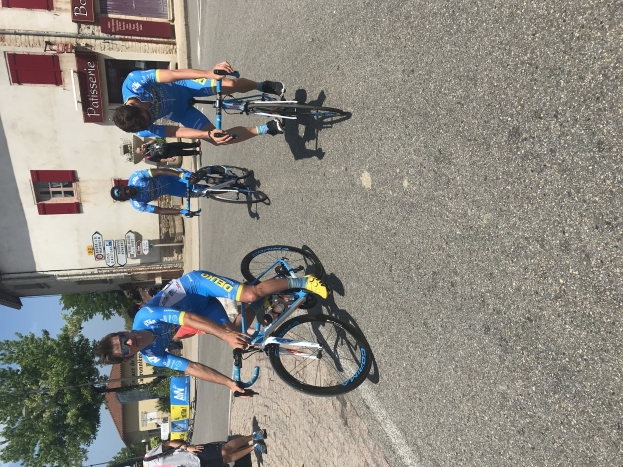 Eine Gruppe von Radfahrern fährt eine Straße entlang an einem Gebäude vorbei, mit Fenstern und Texttafeln, mit Bäumen und einem blauen Himmel im Hintergrund und Zuschauern auf der Straße darunter.