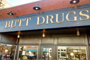 Außensicht eines Gebäudes mit einer beleuchteten Schilder 'butt drugs' und gläserne Wände, die umliegende Fahrzeuge, Gebäude, Bäume und Himmel zeigen.