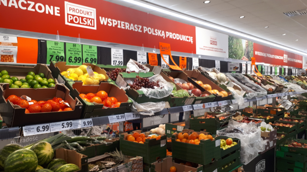 Ladeninneres mit verschiedenen Obstsorten und Gemüse in Kartons mit Preisschildern, Wandtafeln mit Text und Bildern sowie Deckenleuchten.