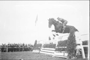 Schwarzes und weißes Foto eines Pferdes und Reiters, die über ein Hindernis springen, bei den 1953 Royal Ascot Horse Trials, mit Zuschauern auf der linken Seite, einer Fahne im Hintergrund und Gras unten.