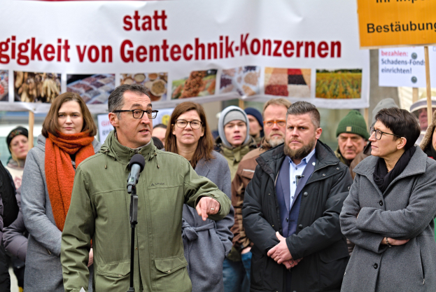 Eine Gruppe von Menschen vor einer Menge mit einem Mann, der in ein Mikrofon spricht, ein Banner mit Text und Bildern dahinter und mehrere Menschen mit Schildern rechts von ihm.
