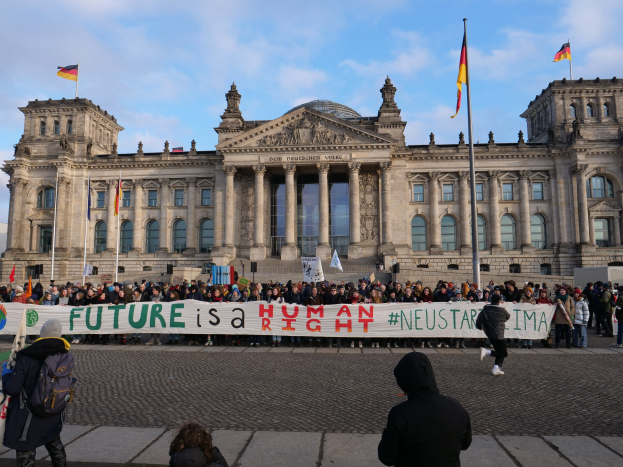Gruppe von Menschen vor dem Reichstagsgebäude in Berlin mit einer Fahne mit der Aufschrift "Zukunft ist ein menschlicher Neustar ima", mit sichtbaren architektonischen Details des Gebäudes und Flaggen im Hintergrund.