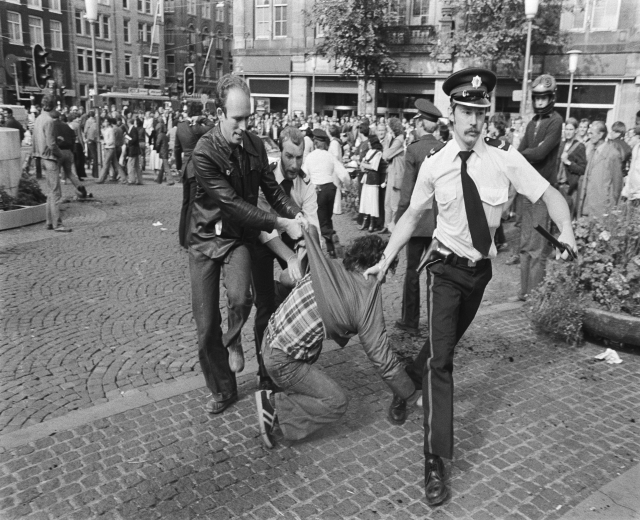Schwarz-weißes Bild eines Mannes, der von zwei Polizisten auf einer Straße mit Zuschauern, Gebäuden, Bäumen und Verkehrsampeln im Hintergrund und einem Mann in Schuhen im Vordergrund gezogen wird.