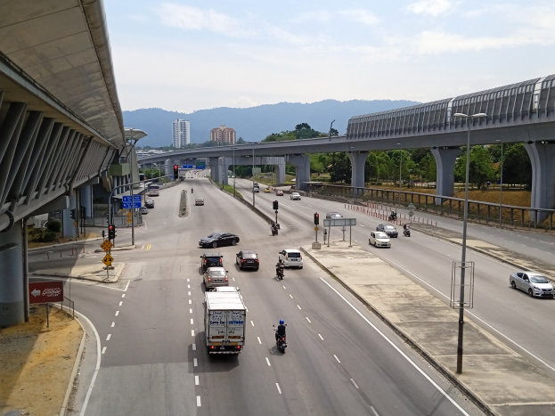 Eine vielbefahrene Autobahn mit mehreren Fahrspuren, neben einer Brücke, mit Straßenlaternen, Ampeln, Verkehrsschildern, Bäumen, Gebäuden, Hügeln und einem bewölkten Himmel im Hintergrund.