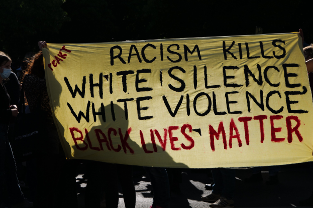 Eine Gruppe maskierter Personen mit einem Banner "Rassismus tötet Stille der Weißen, Gewalt der Weißen, Schwarze Leben zählen" auf einer dunklen Straße.