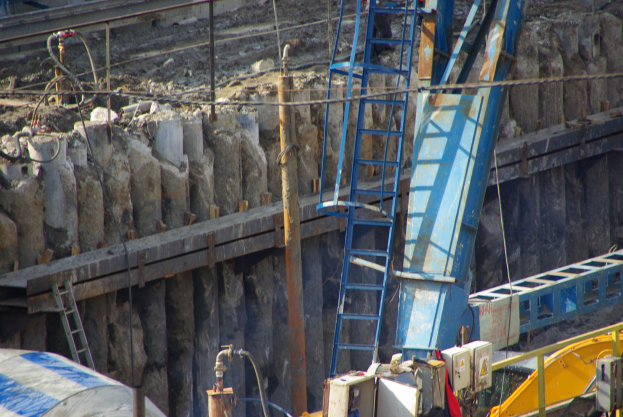Baustelle mit einem großen Kran, Leitern, Stangen, Drähten, Steinen und einem Fahrzeug unten.