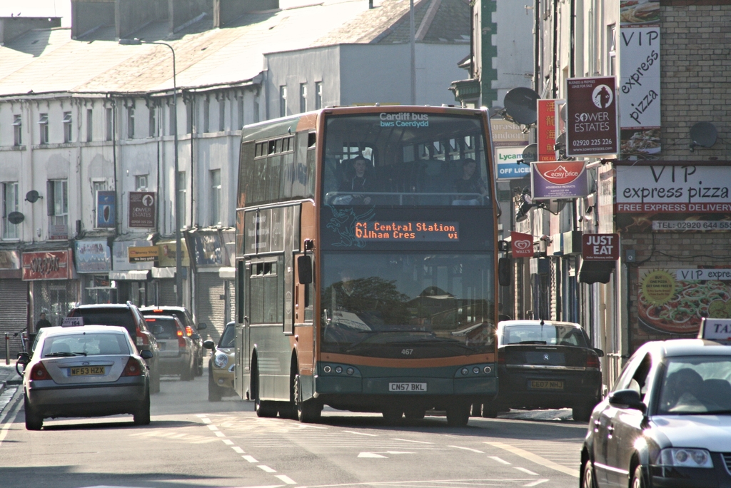 Eine Straße mit Autos und einem Bus im Vordergrund, mit Gebäuden mit Wänden, Fenstern, Tellern und Dächern im Hintergrund, sowie Plakaten und Bannern an den Wänden und einem Pfahl mit einer Straßenlaterne.