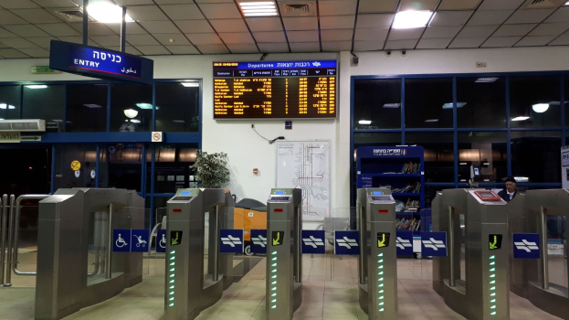 Ein Mann steht an einem Flughafen-Eingang, gegenüber einer Metalldrehtür mit Sicherheitsschleusen, Informationsschildern, einem Topf mit einer Pflanze, einer Wandtafel, einer Decke mit Leuchten und einem Anzeigebildschirm.