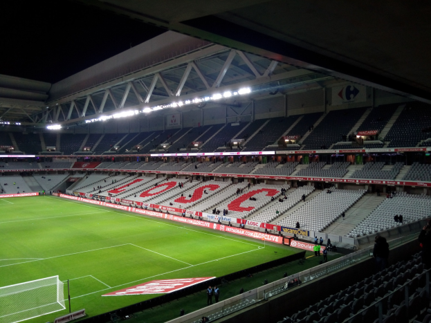 Großes Stadion mit einem grünen Fußballfeld, einem Netz auf der linken Seite, einigen Zuschauern auf den Tribünen und heller Deckenbeleuchtung vor einem dunklen Hintergrund.