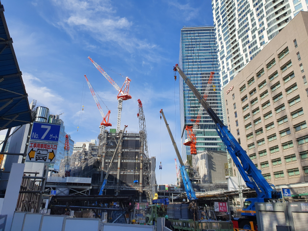 Eine belebte Stadtlandschaft mit hohen Gebäuden, Kränen, Fahrzeugen, Menschen, Schildern, Masten und anderen Gegenständen unter einem bewölktem Himmel.