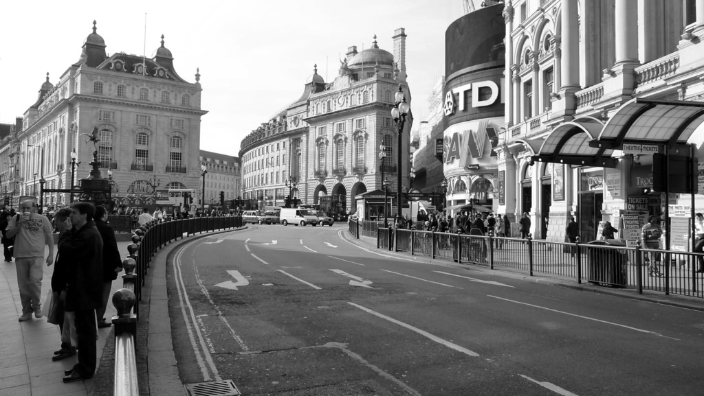Eine Straße mit weißen Linien, Pfeilschildern, einem Geländer auf der rechten Seite und Stahlbarrieren auf der linken Seite, mit Menschen auf beiden Seiten, Straßenlaternen und Gebäuden im Hintergrund.
