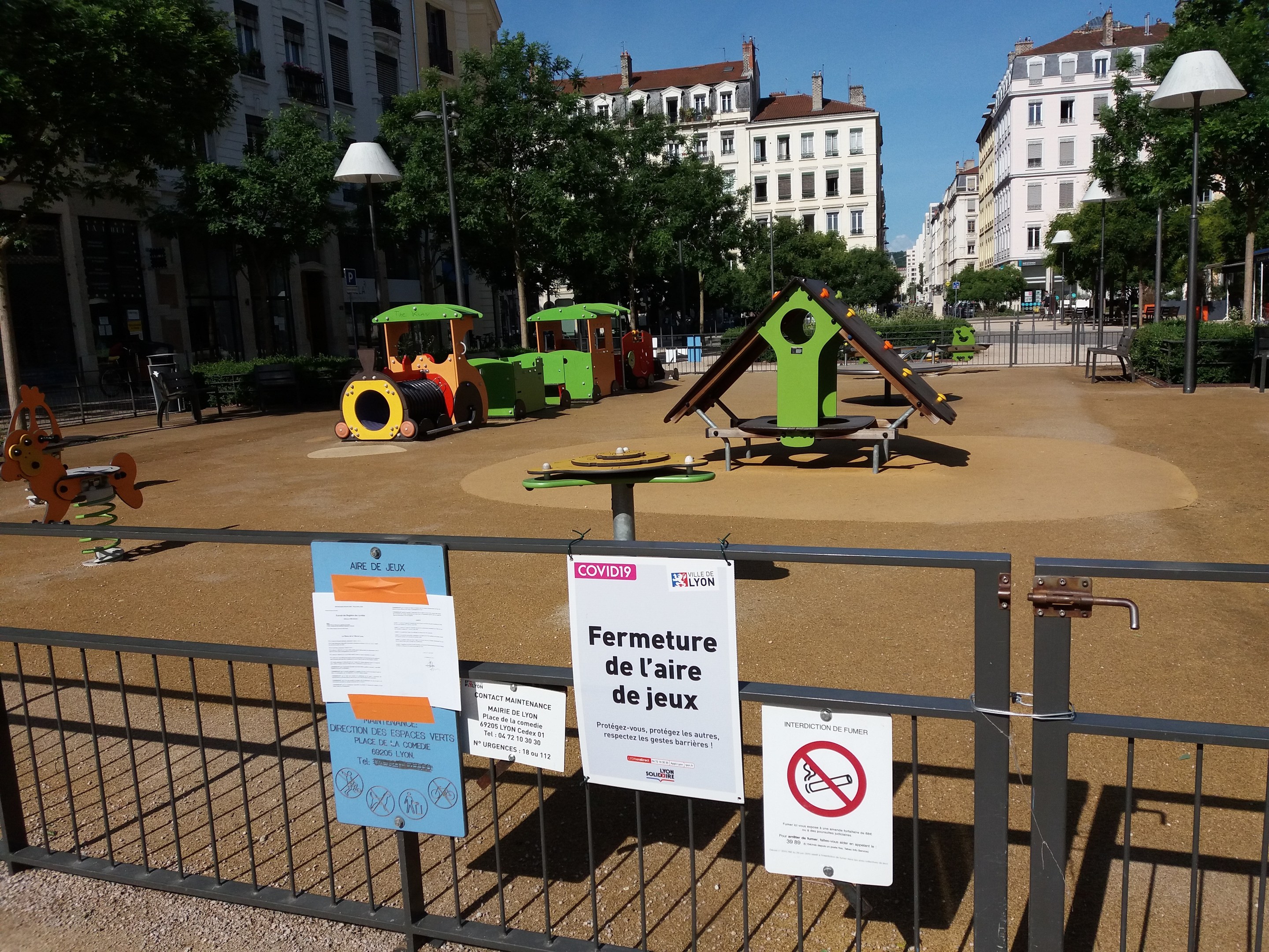 Ein umzäunter städtischer Spielplatz mit mehreren Schildern am Zaun, umgeben von Bäumen, Laternenmasten und Gebäuden unter einem sichtbaren Himmel.