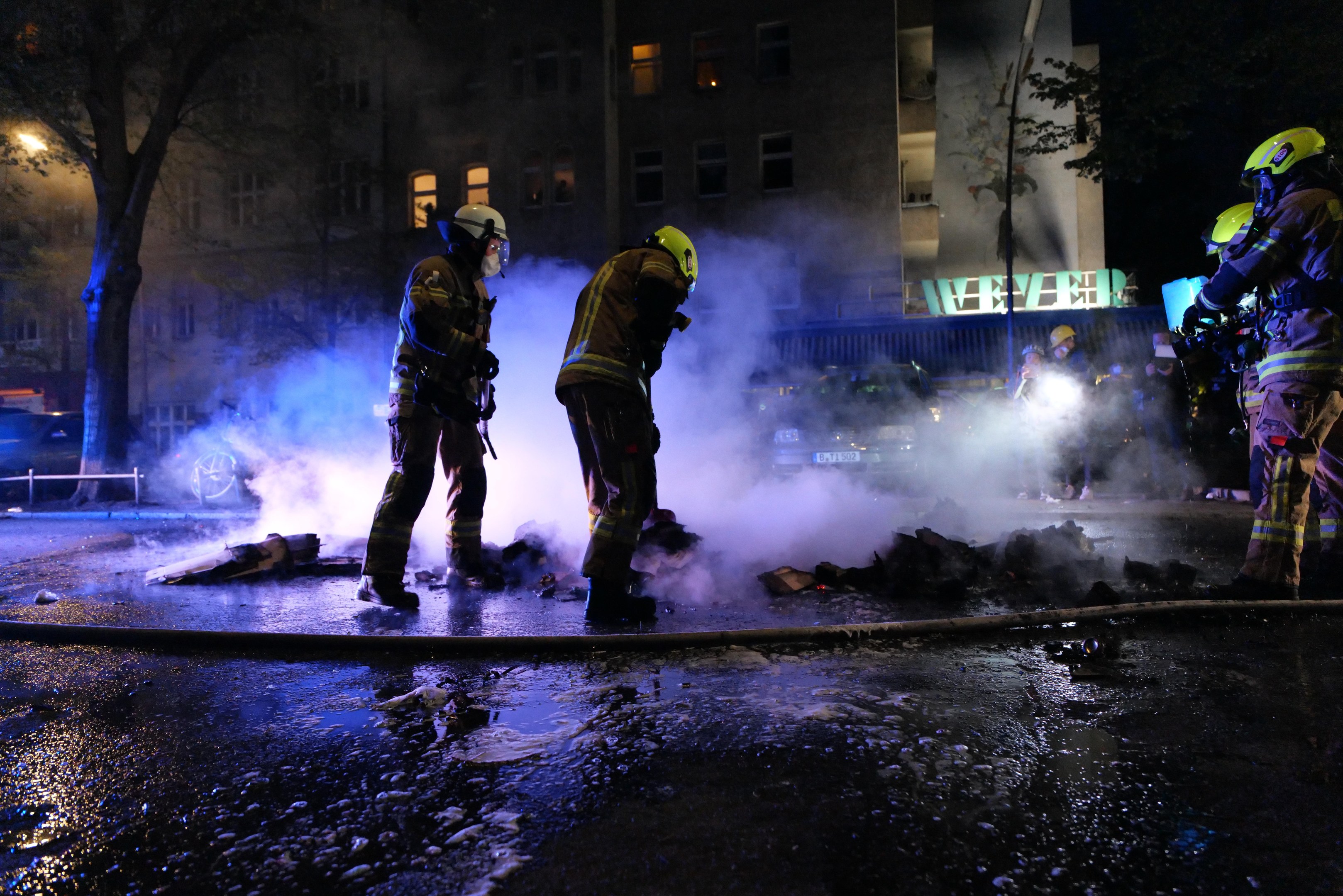 Feuerwehrleute in Helmen stehen nachts auf einer Straße mit Rauch, der aus dem Boden aufsteigt, einem Rohr auf der Straße und Gebäuden, Bäumen und Fahrzeugen im Hintergrund.