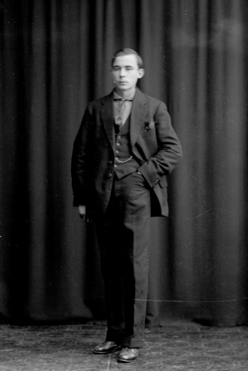 Ein Mann in einem maßgeschneiderten Anzug mit einem strahlend weißen Hemd, gemustertem Schlips und dunklen Hosen steht vor einem Vorhang, sein Haar ist ordentlich frisiert und er hat einen ernsten Gesichtsausdruck.