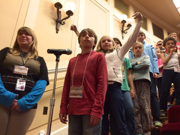Gruppe von Kindern, die vor einem Mikrofon singen, mit einer Frau daneben, Stühlen auf der rechten Seite und beleuchteter Wand im Hintergrund.