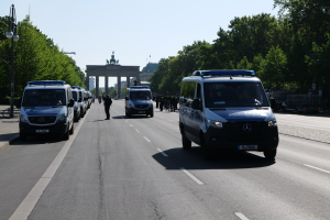 Eine Reihe von Polizeiwagen parkt am Straßenrand vor dem Brandenburger Tor in Berlin. Menschen gehen auf dem Gehweg, Laternenpfähle, Bäume und ein Bogen mit Statuen sind im Hintergrund zu sehen. Der Himmel ist über dem Bogen sichtbar.