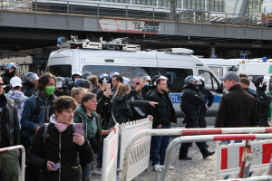 Gruppe von Menschen vor Polizeifahrzeugen mit Barrieren im Vordergrund, einige tragen Helme und halten Telefone, mit einer Brücke und Gebäuden im Hintergrund während einer Demonstration in Berlin, Deutschland.