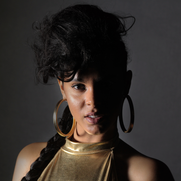 Eine Frau in einem goldenen Kleid und großen Creolen steht selbstbewusst vor einer Wand, ihre glatte Bob-Frisur passt zu ihrem natürlichen Make-up und dem glänzenden Lippenstift und blickt entschlossen in die Kamera.